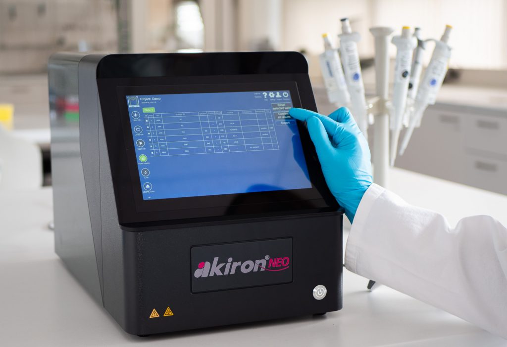 This image shows a piece of laboratory equipment, the akiron NEO, on a clean countertop. A person wearing a white lab coat and blue gloves is using their index finger to interact with the device's touchscreen. In the background, there is a rack of pipettes, confirming the laboratory setting. The device's screen displays a table of data, suggesting it's used for analysis or testing.