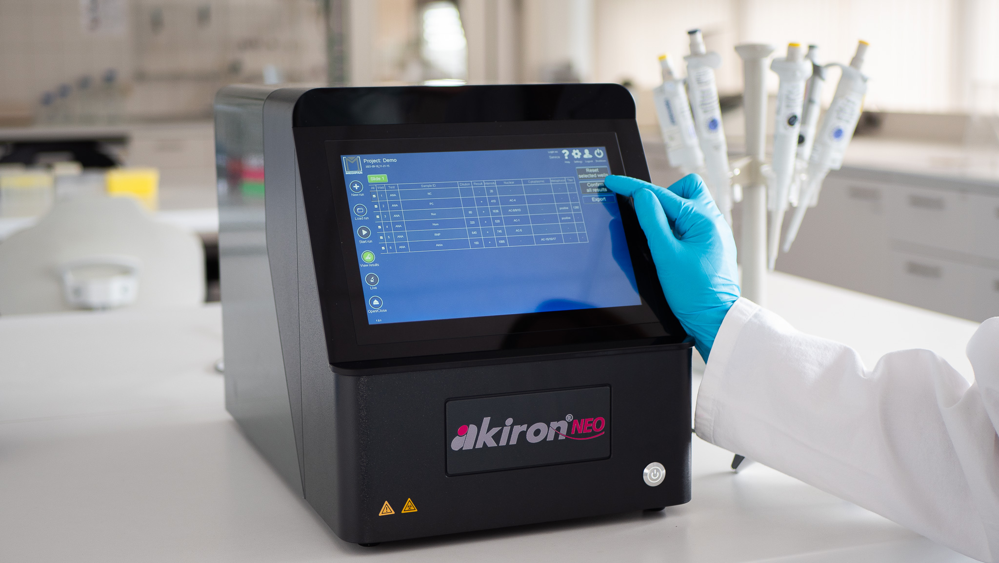 This image shows a piece of laboratory equipment, the akiron NEO, on a clean countertop. A person wearing a white lab coat and blue gloves is using their index finger to interact with the device's touchscreen. In the background, there is a rack of pipettes, confirming the laboratory setting. The device's screen displays a table of data, suggesting it's used for analysis or testing.