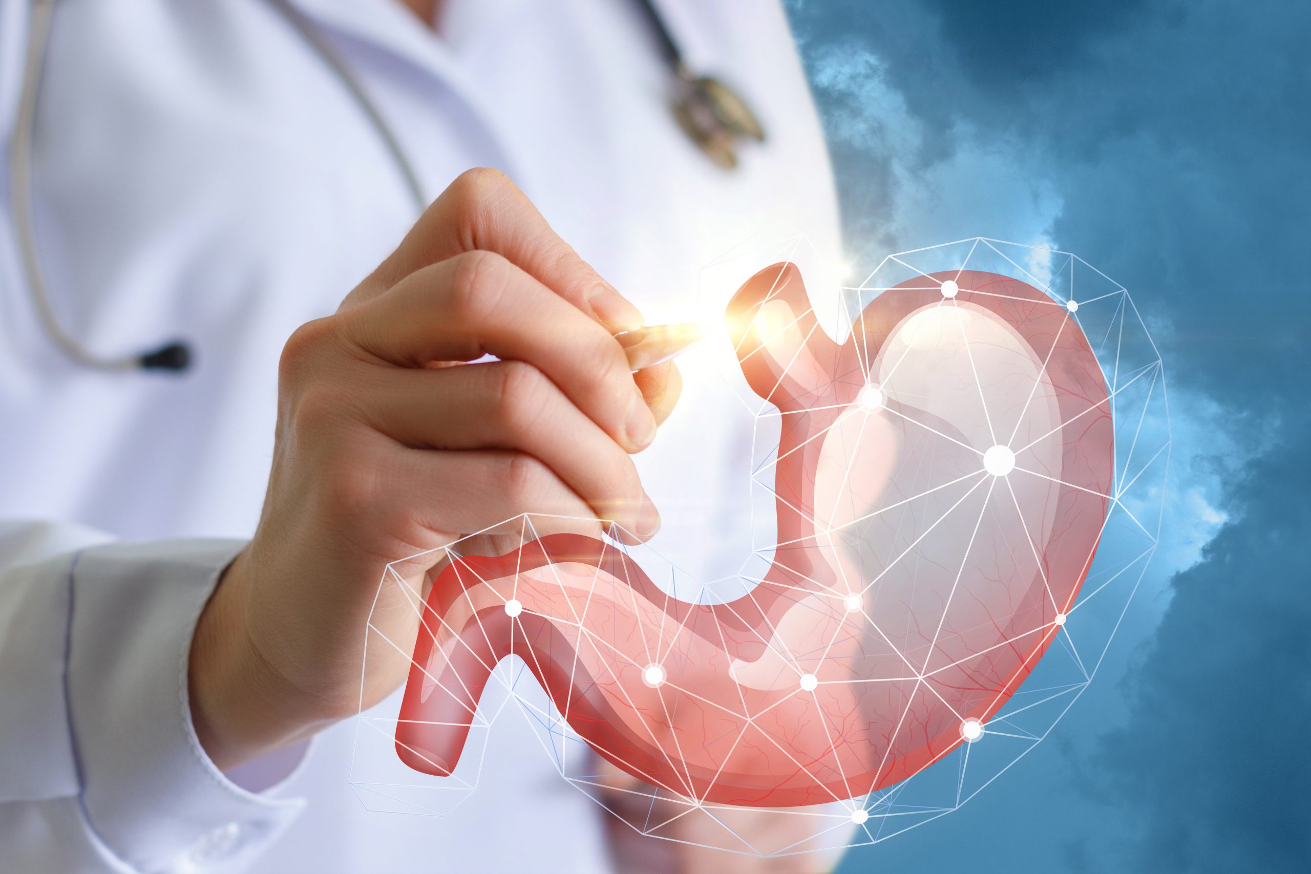 The image shows a medical professional, likely a doctor, holding a digital illustration of a human stomach. The stomach appears semi-transparent with visible blood vessels, and it is overlaid with a network of white lines and dots, giving it a high-tech, futuristic appearance. The background features a cloudy sky, adding a sense of openness and highlighting the digital and medical theme. The doctor is wearing a white coat and has a stethoscope around their neck.