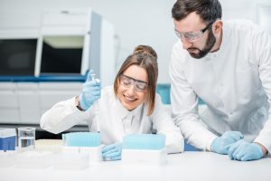 This image shows a man and a woman in a lab, both wearing lab coats and safety glasses. The woman is smiling as she uses a pipette, while the man looks on.