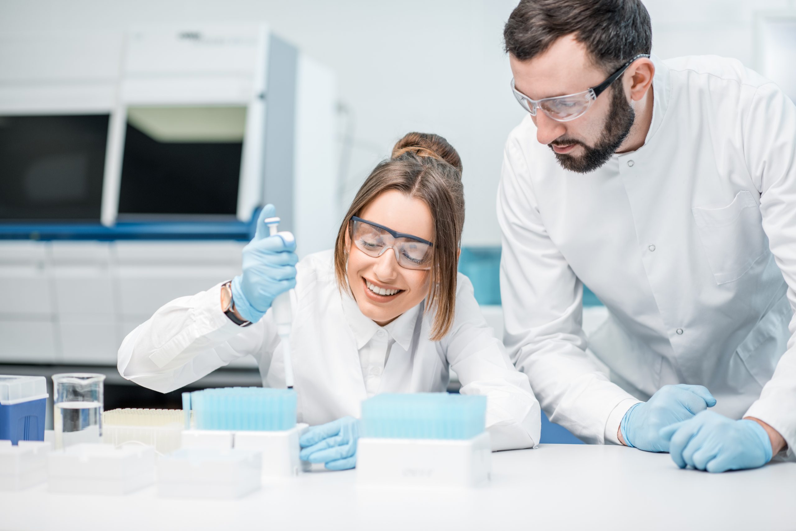 This image shows a man and a woman in a lab, both wearing lab coats and safety glasses. The woman is smiling as she uses a pipette, while the man looks on.