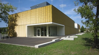 This image shows a modern, two-story building with a clean, cubic architecture. The upper half of the facade is covered with gold or brass-colored metal panels. The lower part of the building has a light, simple cladding that continues into a covered, recessed entrance area with large glass windows. The building is located on a well-maintained lawn with some trees in the background and on the sides. An asphalted area leads to the main entrance.