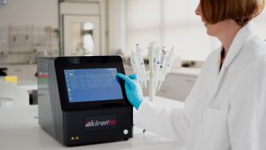 A person wearing a white lab coat and blue gloves is interacting with the akiron® NEO device's touchscreen interface. The screen shows a data table, suggesting the machine is used for analysis or diagnostics. In the background, other lab equipment, including a stand with multiple pipettes, is visible, confirming the setting is a scientific or medical laboratory.