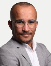 The image is a professional headshot of a man.
He is wearing a light grey blazer over a white shirt, and a faint grey pattern is visible on the blazer. He has a neatly trimmed beard and mustache, and is wearing thin-rimmed glasses. The man is looking directly at the camera with a slight smile. The background is a solid, light grey.