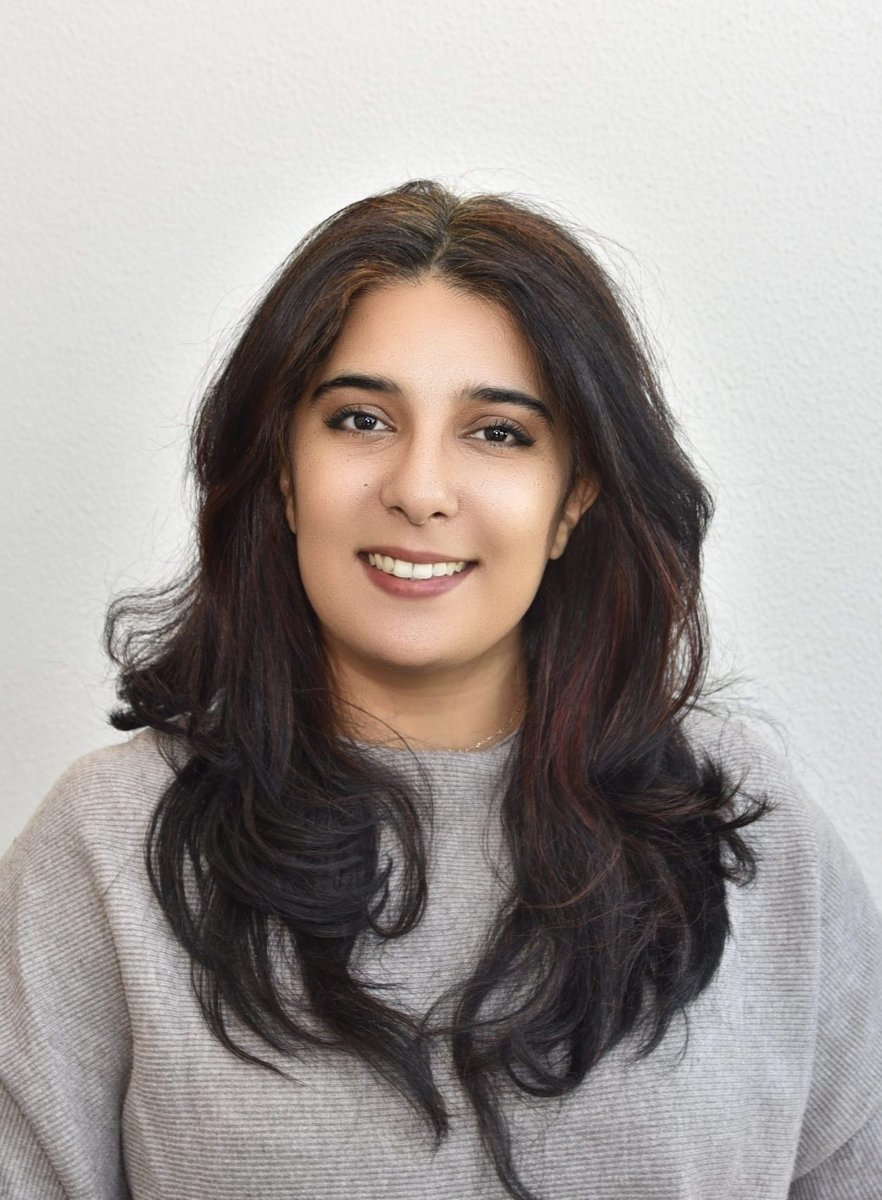 The image shows a smiling woman with long, dark, wavy hair. She is wearing a light-colored, possibly grey, knitted sweater. The background is a simple, light wall. She is looking directly at the camera.