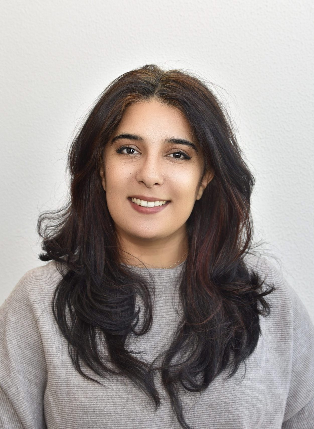 Anfrage erfolgreich

The image shows a smiling woman with long, dark, wavy hair. She is wearing a light-colored, possibly grey, knitted sweater. The background is a simple, light wall. She is looking directly at the camera.