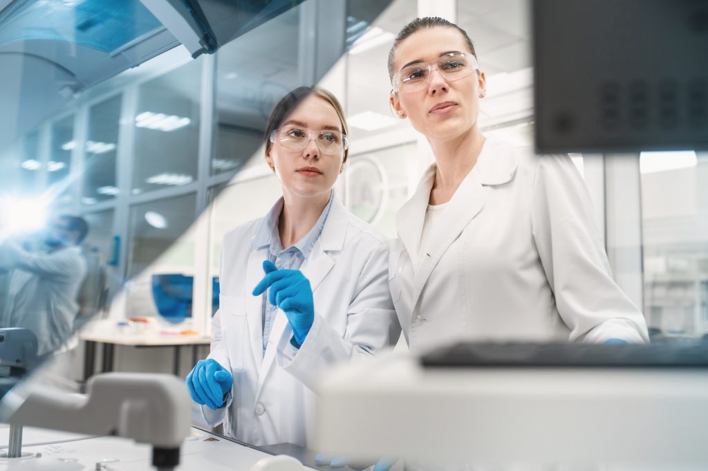 Two women are visible in the image, standing in a lab and wearing white lab coats and safety glasses. They appear to be working on a scientific project or experiment, looking intently at a computer monitor. The woman on the left is also wearing blue latex gloves and is pointing at something with her finger.In the background, other lab equipment and another person who is also working can be seen.