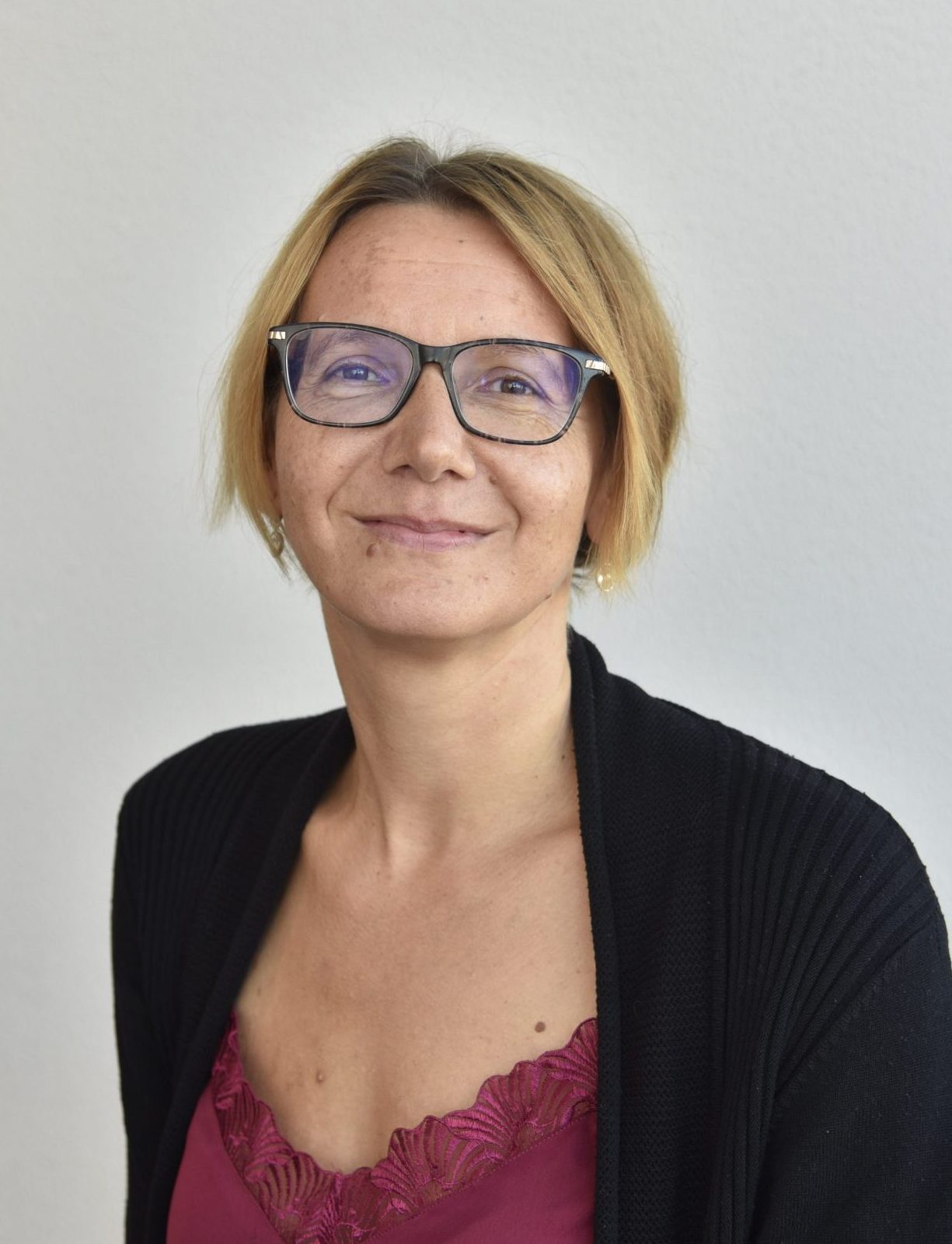 The image shows a woman with short, blonde hair and glasses. She is wearing a black cardigan over a dark red top. She is smiling slightly and looking at the camera. The background is a plain, light-colored wall.