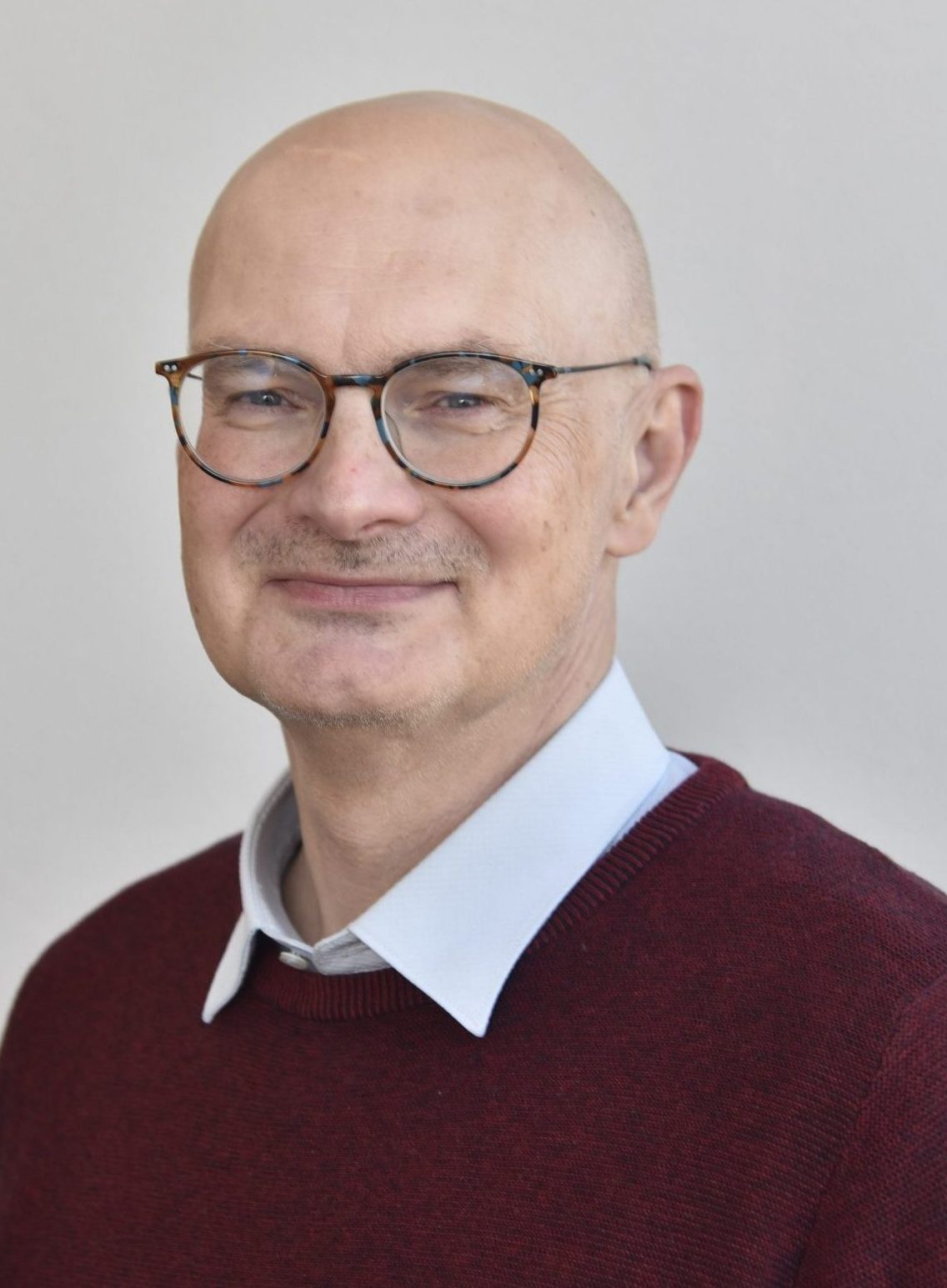 The picture shows a head-and-shoulders portrait of Prof. Dr. Dirk Roggenbuck is the CEO of both Medipan and GA Generic Assays. He has a bald head,in his late 50s or 60s and is wearing round-framed glasses. He is smiling slightly, with a friendly and approachable expression. He is dressed in a dark red sweater over a light blue collared shirt. The background is a plain, light-colored wall. The lighting is soft and even, highlighting his face.