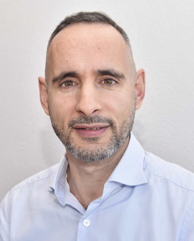 Middle-aged man with short, dark hair that is heavily trimmed at the sides. He has a well-groomed, short beard that contains some gray hairs. His eyes are brown, and he looks directly into the camera, giving a friendly and open impression.

He is wearing a light blue shirt that is neatly ironed, with the top button open, giving him a slightly relaxed but professional look. The background is plain and light, probably a white or very light-colored wall, which makes the person stand out clearly in the foreground.

Translated with DeepL.com (free version)