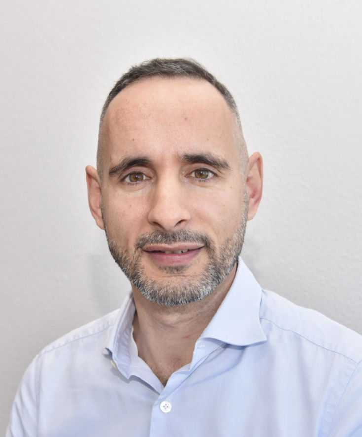 Middle-aged man with short, dark hair that is heavily trimmed at the sides. He has a well-groomed, short beard that contains some gray hairs. His eyes are brown, and he looks directly into the camera, giving a friendly and open impression.

He is wearing a light blue shirt that is neatly ironed, with the top button open, giving him a slightly relaxed but professional look. The background is plain and light, probably a white or very light-colored wall, which makes the person stand out clearly in the foreground.

Translated with DeepL.com (free version)