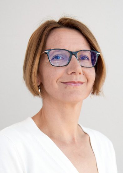 This is a portrait of a woman with short, light brown hair and fair skin. She is wearing glasses with dark frames and a white top. The background is plain and light-colored, which keeps the focus on her face. She has a slight, confident smile and is looking directly at the camera.