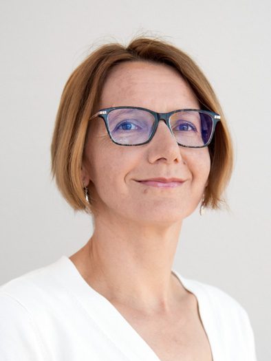 This is a portrait of a woman with short, light brown hair and fair skin. She is wearing glasses with dark frames and a white top. The background is plain and light-colored, which keeps the focus on her face. She has a slight, confident smile and is looking directly at the camera.