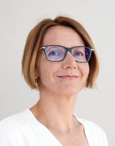 Dies ist ein Porträt einer Frau mit kurzen, hellbraunen Haaren und heller Haut. Sie trägt eine Brille mit dunklem Gestell und ein weißes Oberteil. Der Hintergrund ist schlicht und hell, sodass der Fokus auf ihrem Gesicht bleibt. Sie lächelt leicht selbstbewusst und blickt direkt in die Kamer