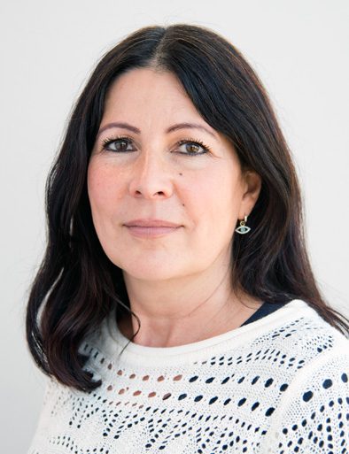 This is a portrait of a woman with shoulder-length, dark brown hair. She has a fair complexion and is wearing subtle makeup. She is looking directly at the camera with a calm and confident expression. She is dressed in a white, knitted top with a pattern of small holes