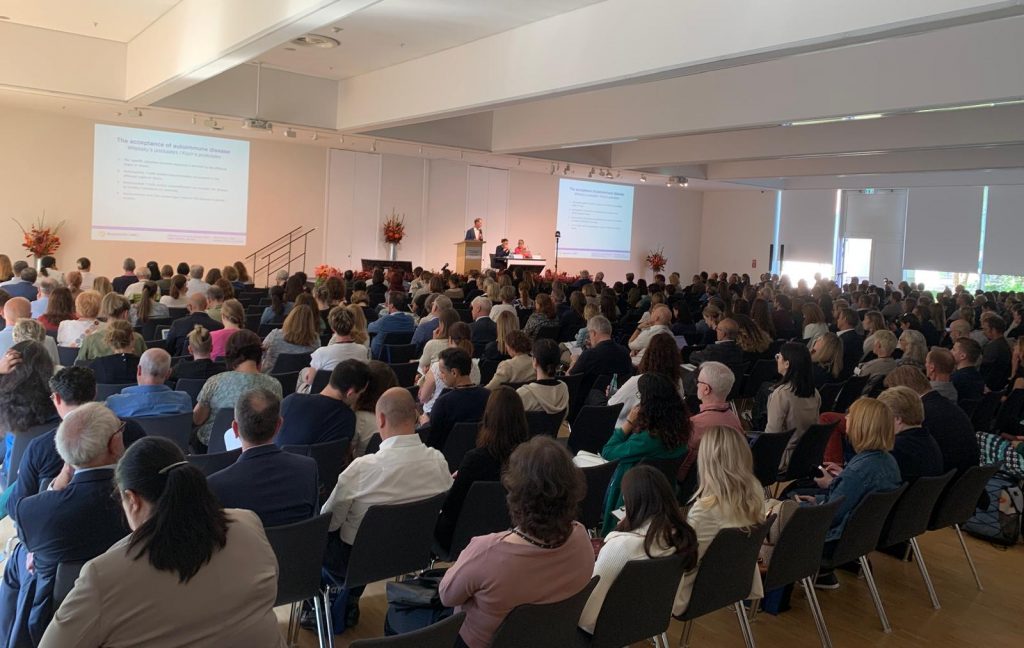 The image shows a man giving a presentation ath the From Biomarkers to AI: Highlights of the 17th Dresden Symposium on Autoantibodies 2025. He is standing near a large screen displaying a slide titled "Planned IIFT Workflow" with a diagram and some text. The audience is seated and attentively listening, with some taking notes. There are flowers on a stand next to the presenter, and a grand piano is also visible in the room. The setting appears professional and focused on a technical or scientific topic.