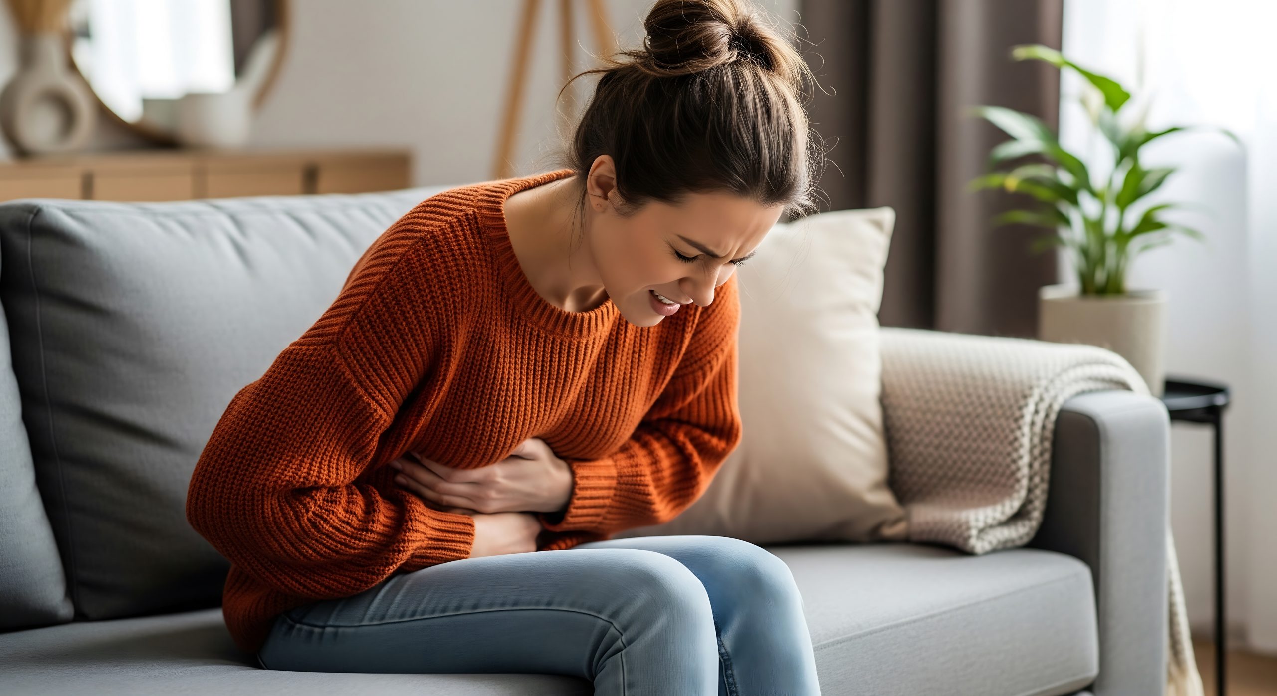 A young woman sits on a grey sofa, leaning forward and clutching her stomach with an expression of pain and discomfort.