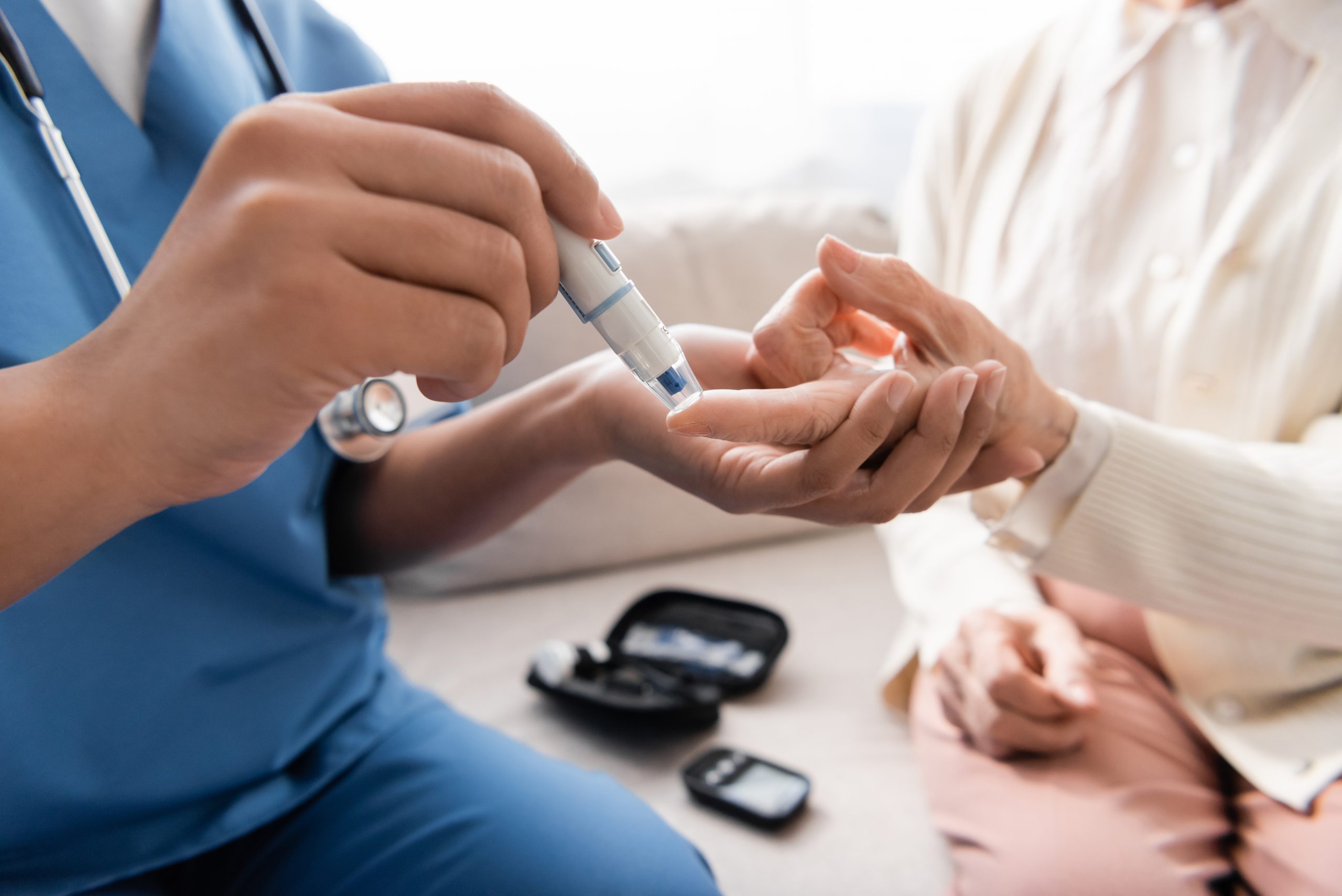 A healthcare professional in blue scrubs pricks the finger of a patient using a lancing device to check their blood sugar levels. A glucose meter and testing kit are visible on the seat nearby.