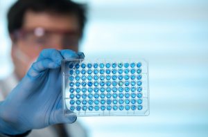 A Researcher wearing protective gear and blue gloves holds a 96-well microplate filled with a blue substance, highlighting a single greenish-blue outlier well against a blurred laboratory background.